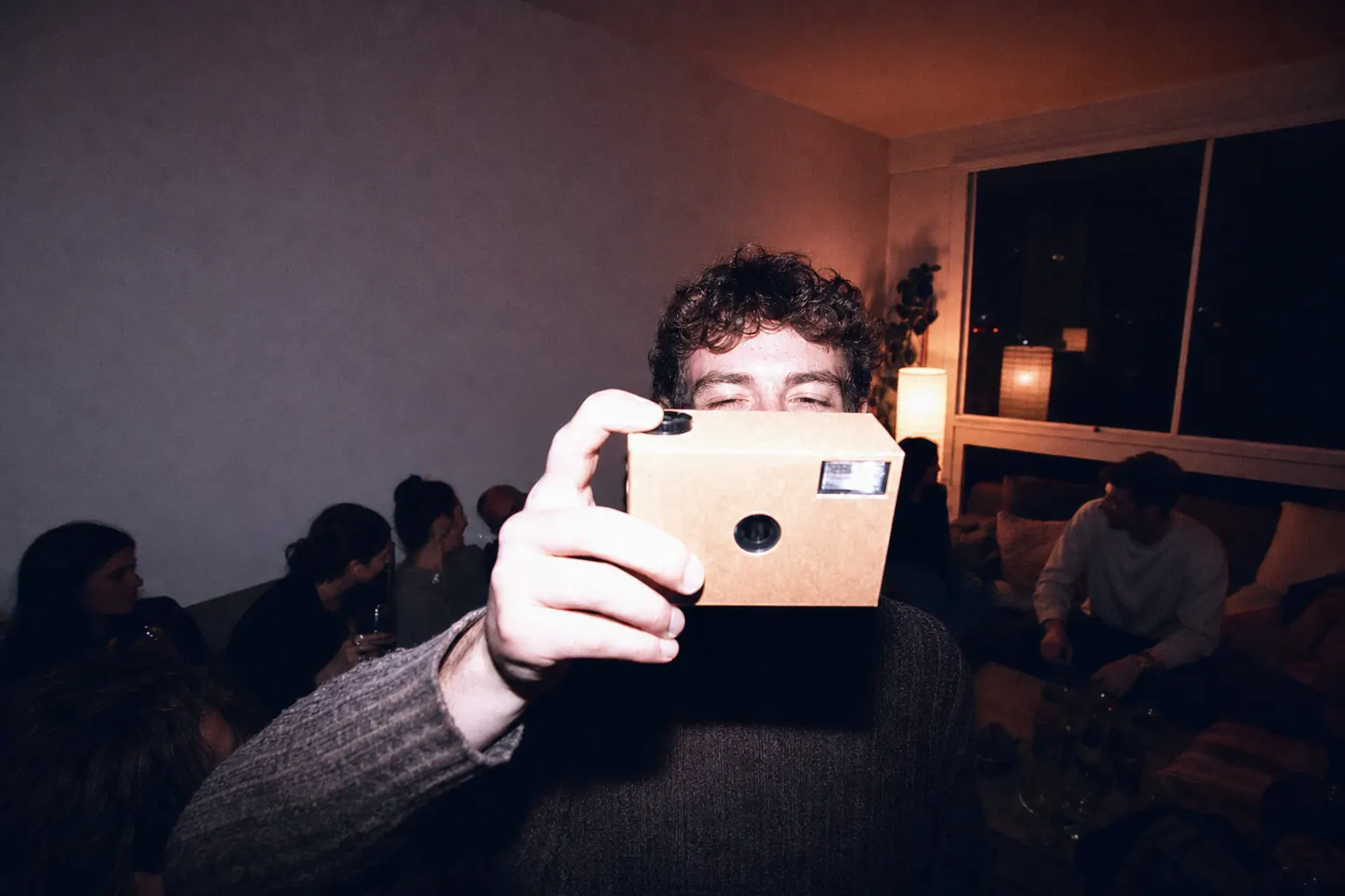 A plain cardboard-bodied single-use camera held up at a dim night gathering, flash lighting the hand while the room falls into shadow